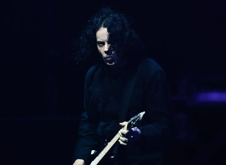 Guitarist with long dark hair in black clothing playing an electric guitar on a dimly lit stage, focused on the instrument.