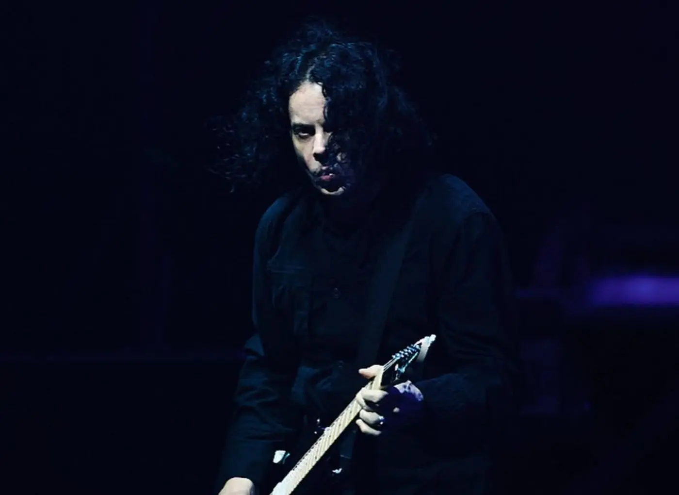 Guitarist with long dark hair in black clothing playing an electric guitar on a dimly lit stage, focused on the instrument.