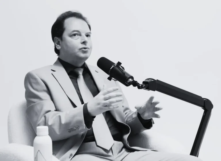 Man in a light suit and tie sits in a chair, speaking into a mounted microphone during a podcast or interview session.