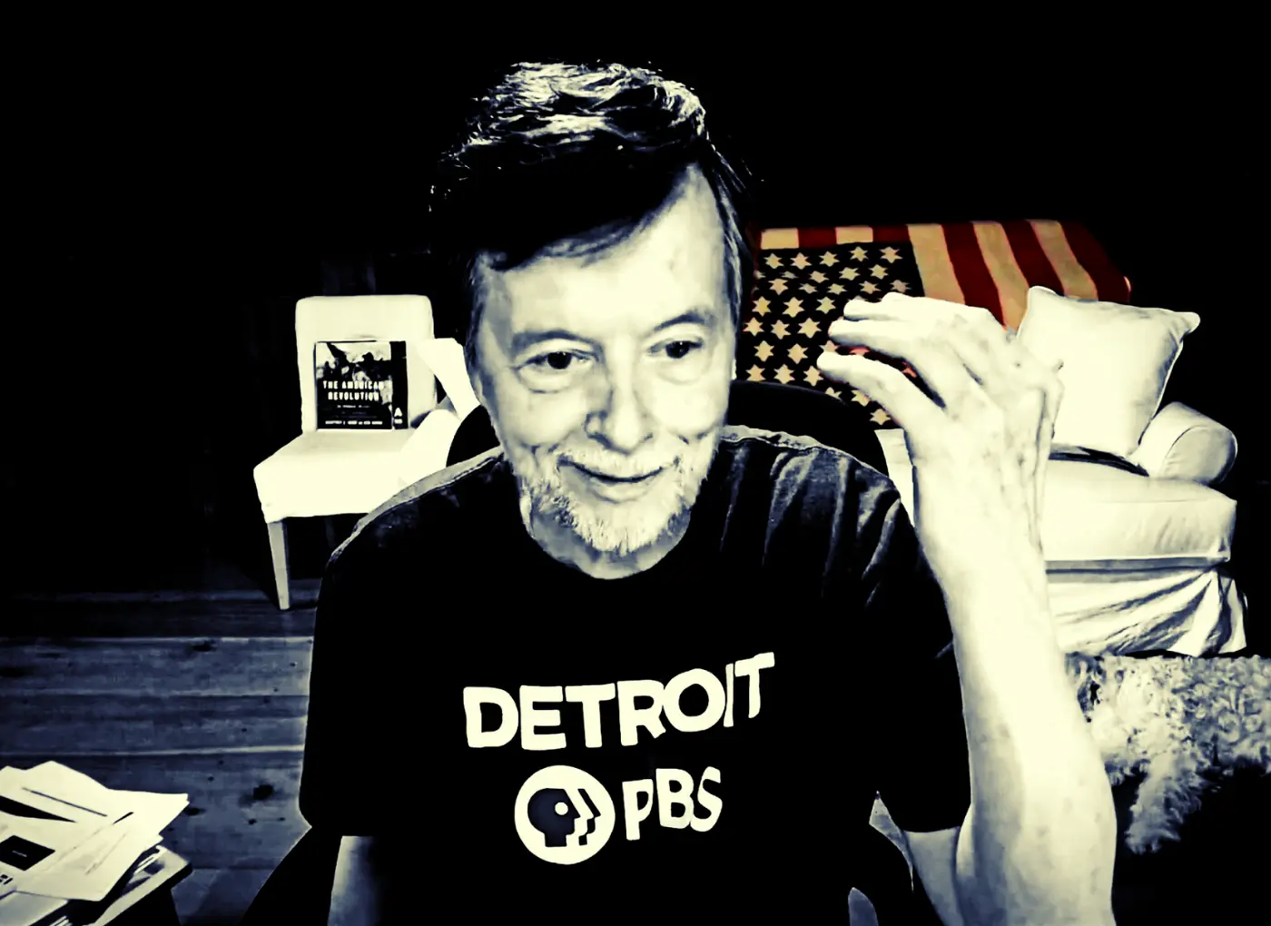 Smiling man with a gray beard wearing a Detroit PBS T-shirt, seated in a room with an American flag blanket on a sofa behind him.