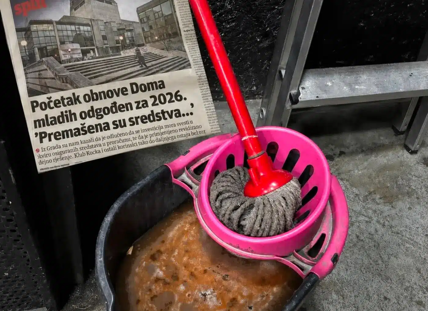 Newspaper with a bold headline about delaying the youth home renovation to 2026, placed next to a pink mop wringer and mop in a dirty utility area.