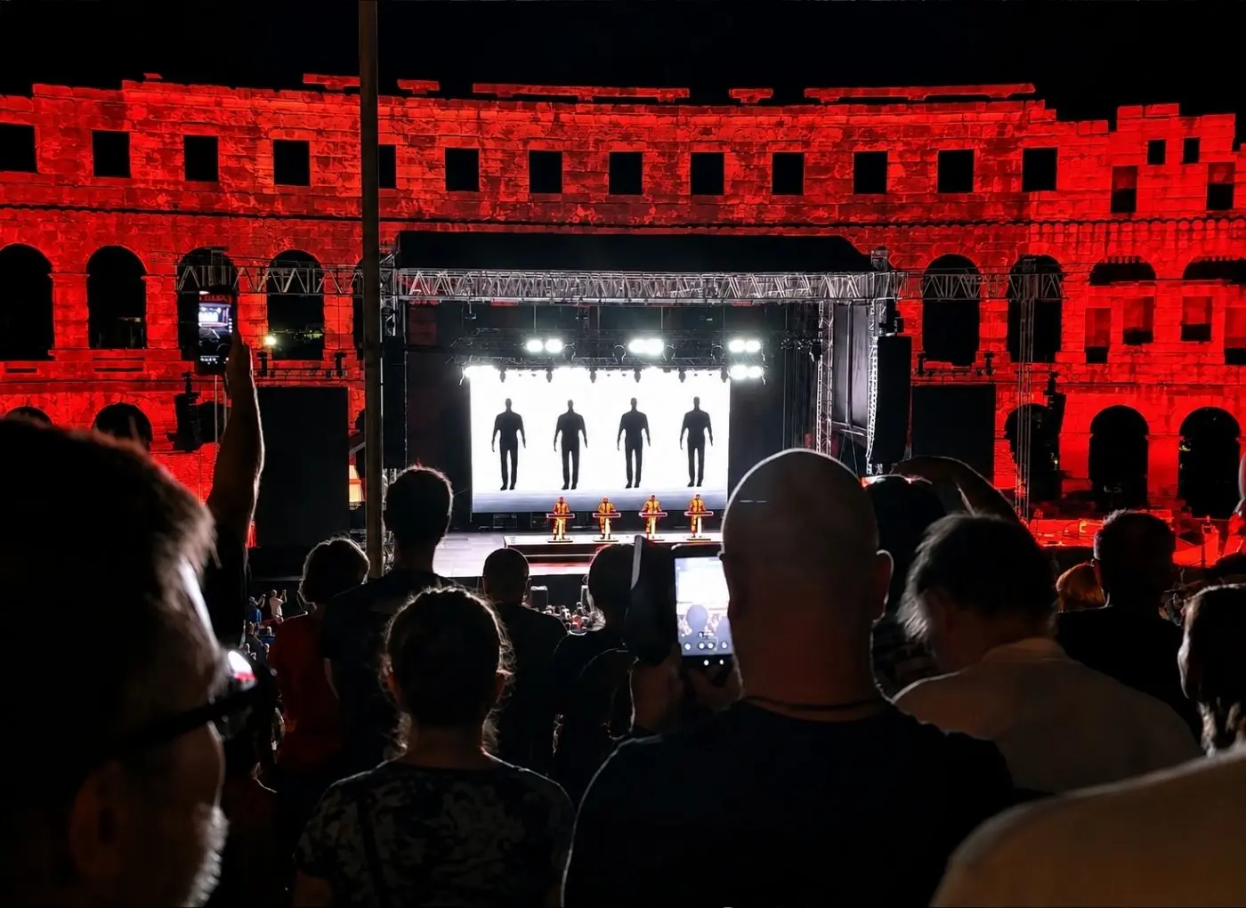 Crowd watches a live performance at night in a red-lit ancient amphitheater, with five silhouetted performers on a bright stage.