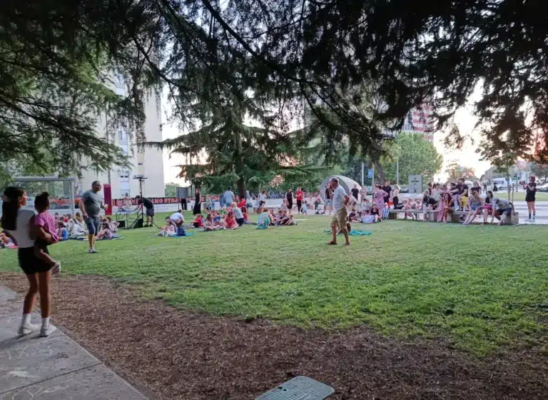 Group of people sitting on blankets in a city park during an outdoor gathering under trees.