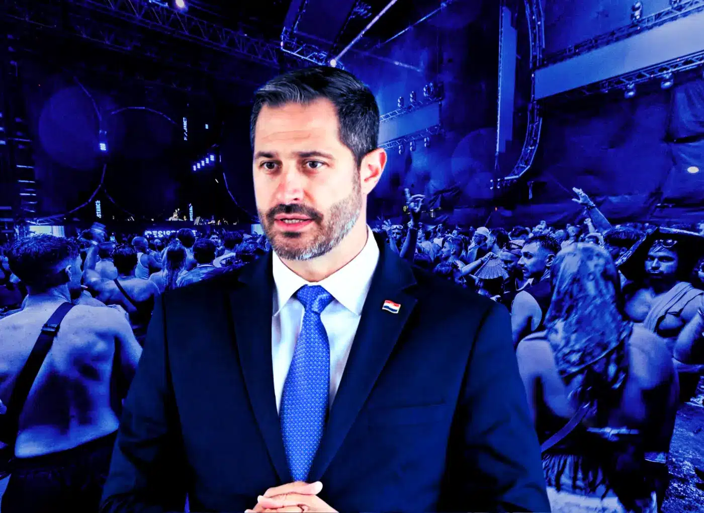 Man in a dark suit and blue tie stands in front of a large blue-lit concert crowd.