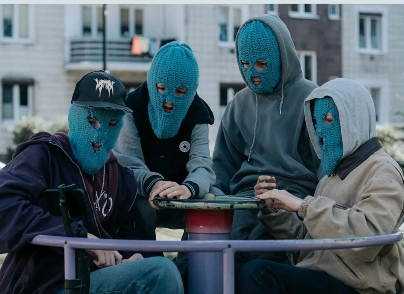 Four people wearing blue knit balaclavas huddle around a round playground spinner in a park or courtyard.