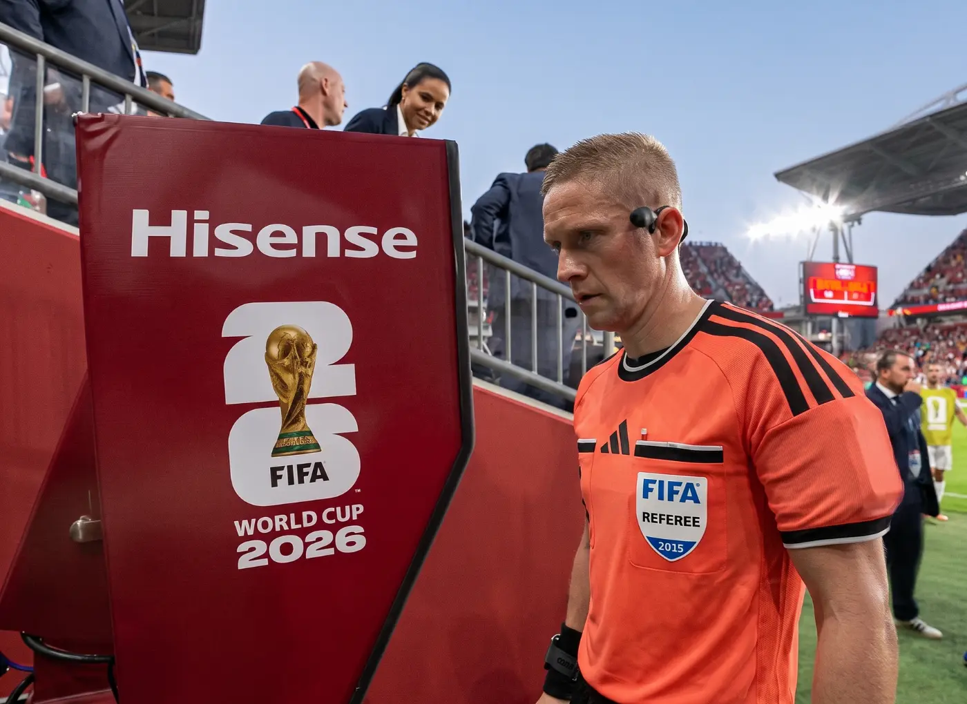 Referee in an orange uniform with an earpiece stands beside a red barrier displaying the FIFA World Cup 2026 logo in a stadium.