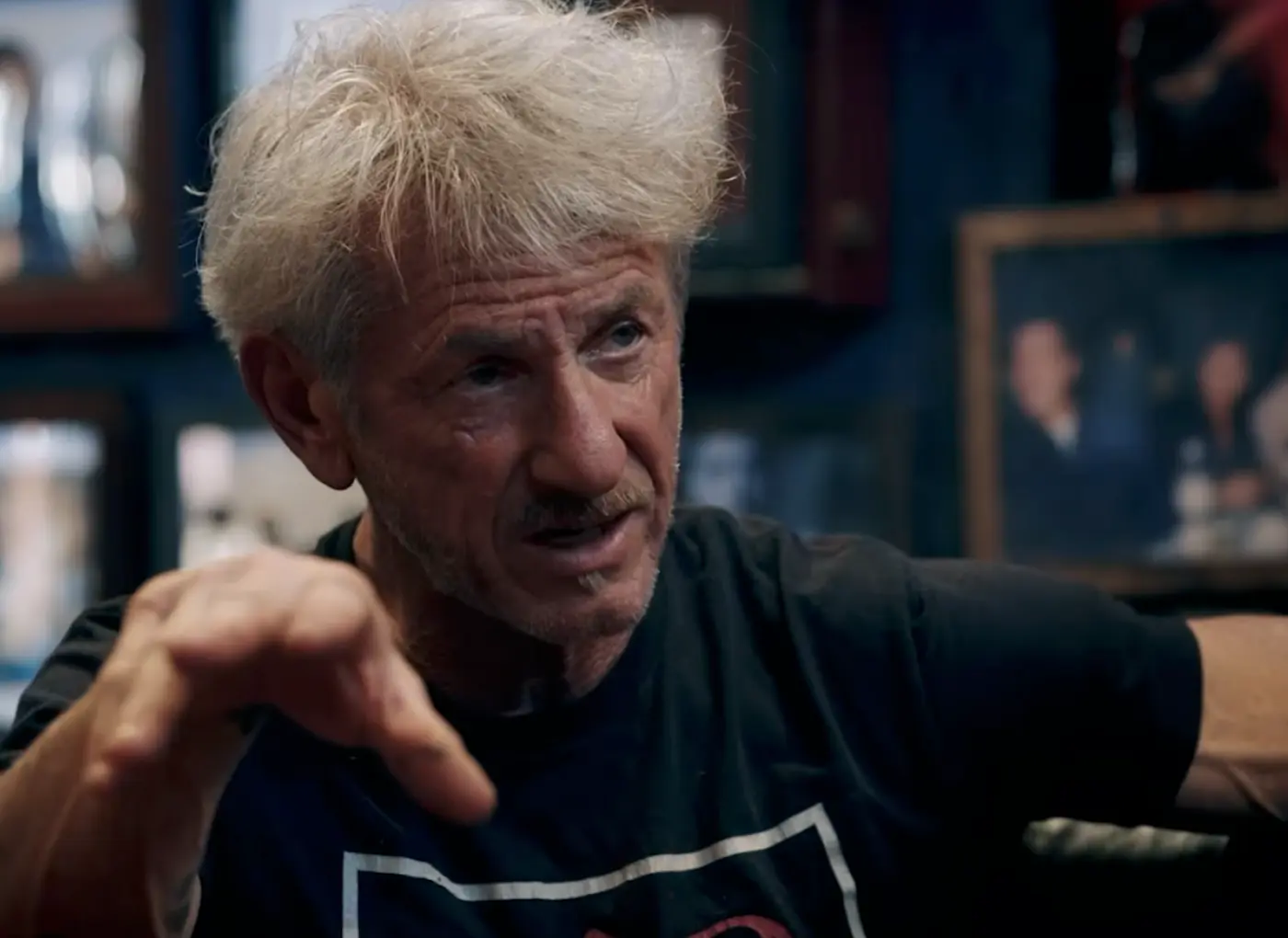 Older man with gray hair and stubble, wearing a black shirt, points as he speaks in a dim room with framed photos in the background.