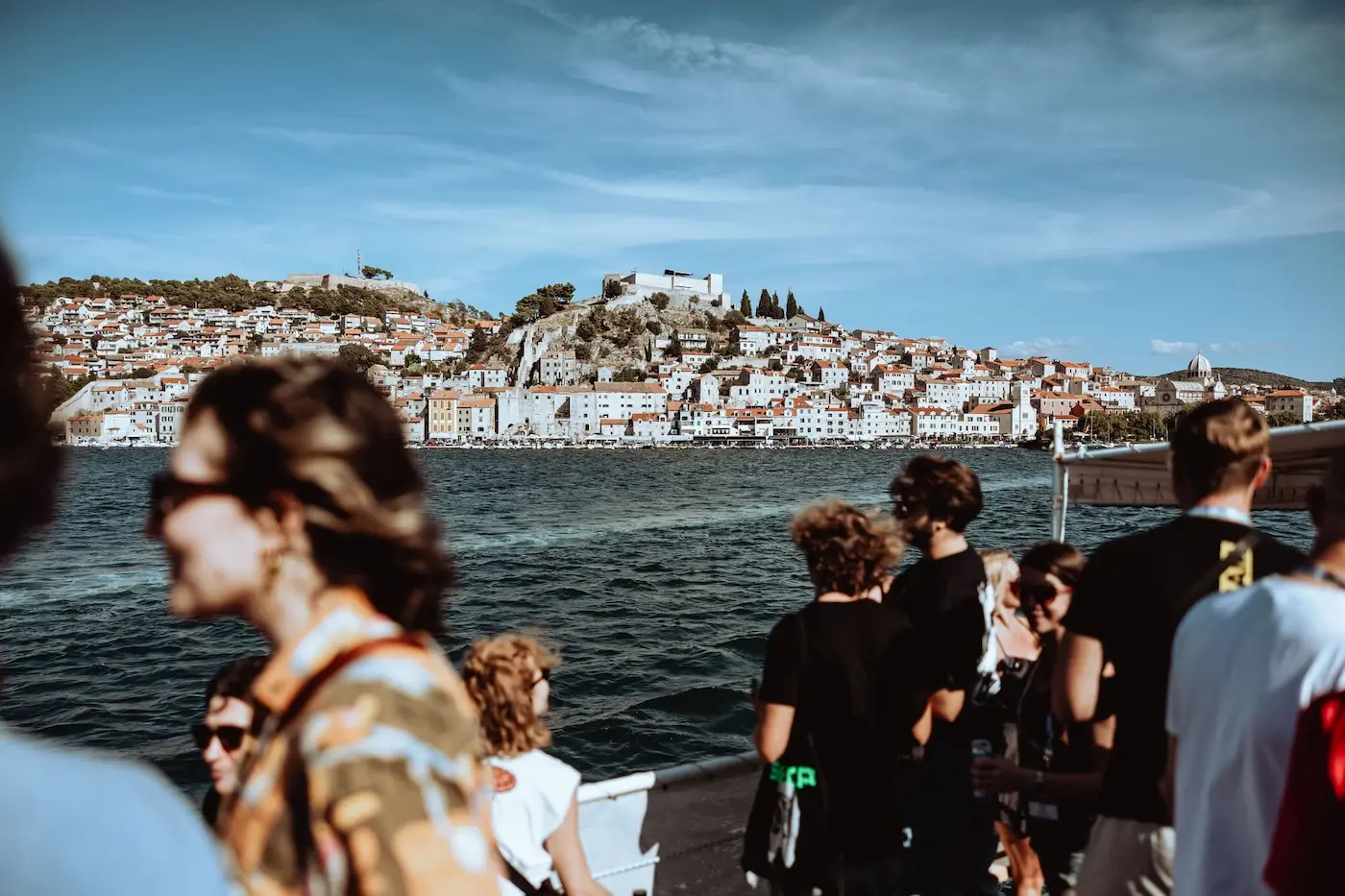 Crowded ferry deck with tourists looking toward a hillside town across the blue bay.