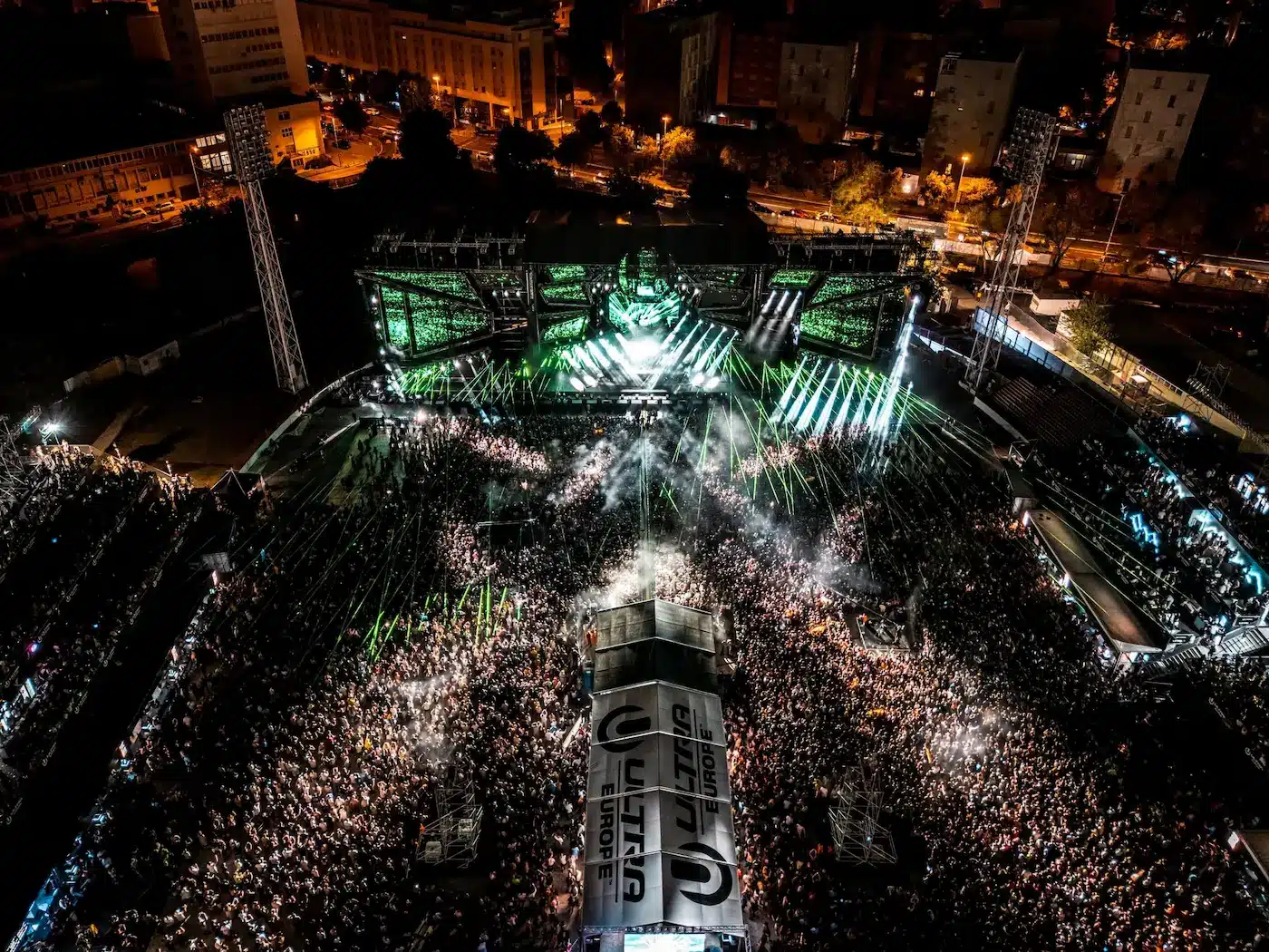Aerial view of a large nighttime outdoor concert with a bright central stage, green laser beams, and a dense crowd.