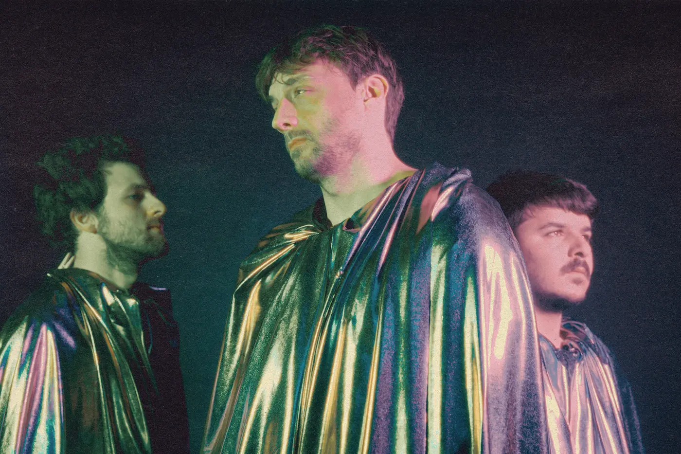 Three men in iridescent metallic capes stand against a dark background, heads turned in profile, a moody, artistic portrait of a band.