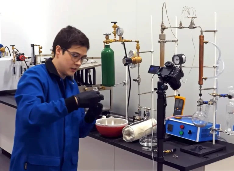 Lab technician in a blue coat and gloves setting up glassware near a gas manifold in a chemistry lab.