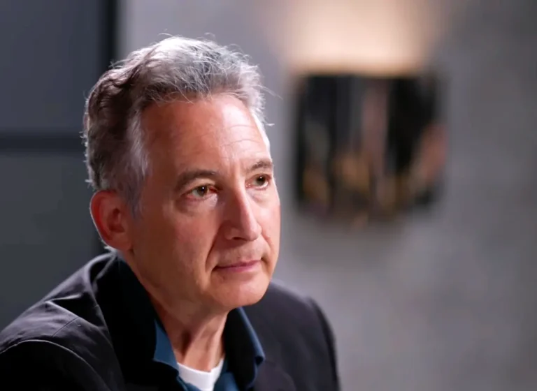 Close-up of a middle-aged man with gray hair in a dark jacket, looking thoughtful during an interview.