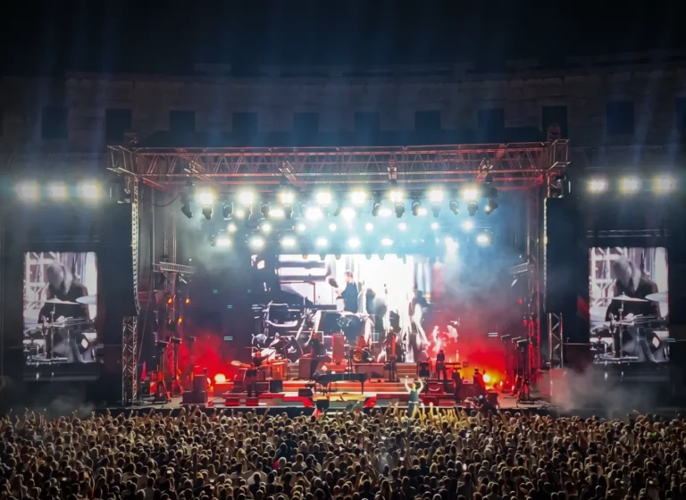 Outdoor concert at night with a brightly lit stage, smoke, and a dense cheering crowd in front of it.