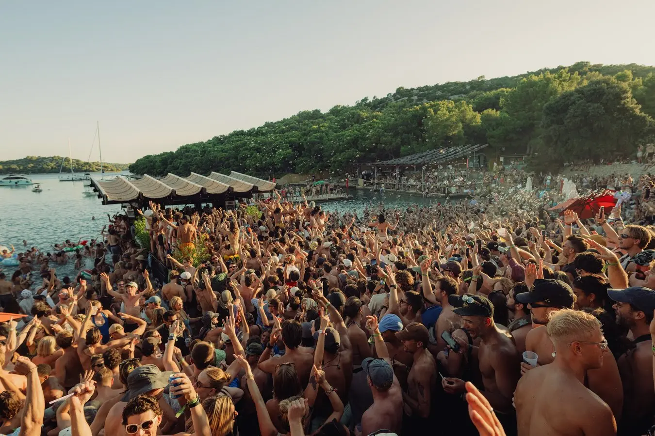 Large crowd of people cheering with hands raised at a seaside party on a dock and beach at sunset. Boats in the water in the background.
