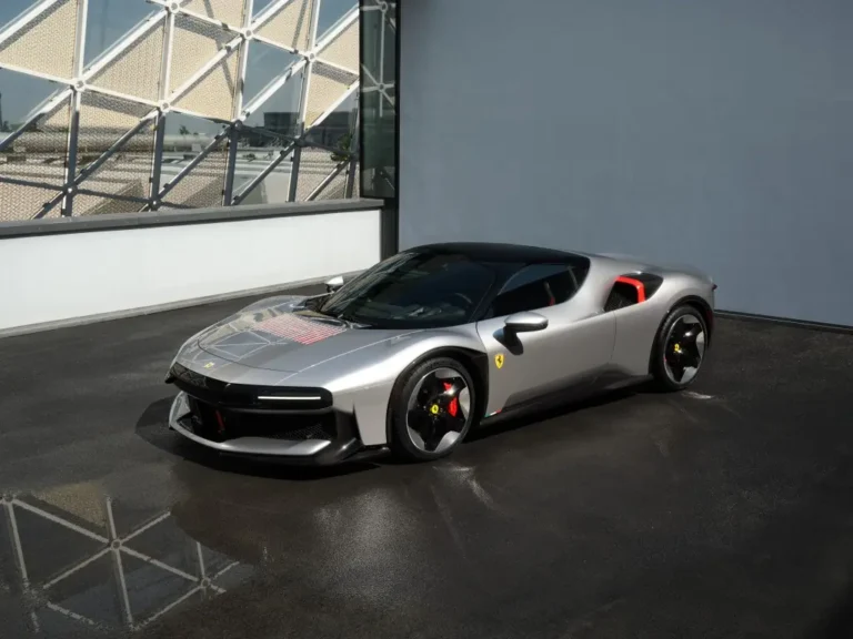 Front three-quarter view of a metallic silver Ferrari sports car with black roof and red interior, parked on a dark pavement beside a modern glass wall.