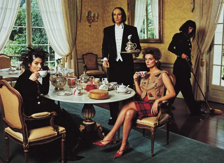 Elegant dining room scene with two women seated at a teatable, a waiter serving tea, and a maid sweeping in the background.