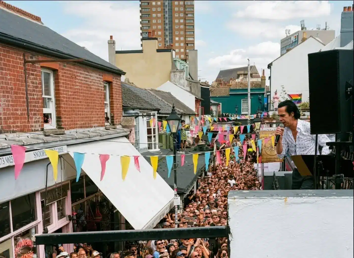 Male singer performs on a rooftop stage above a crowded street festival with colorful bunting flags strung across the street in a town setting.