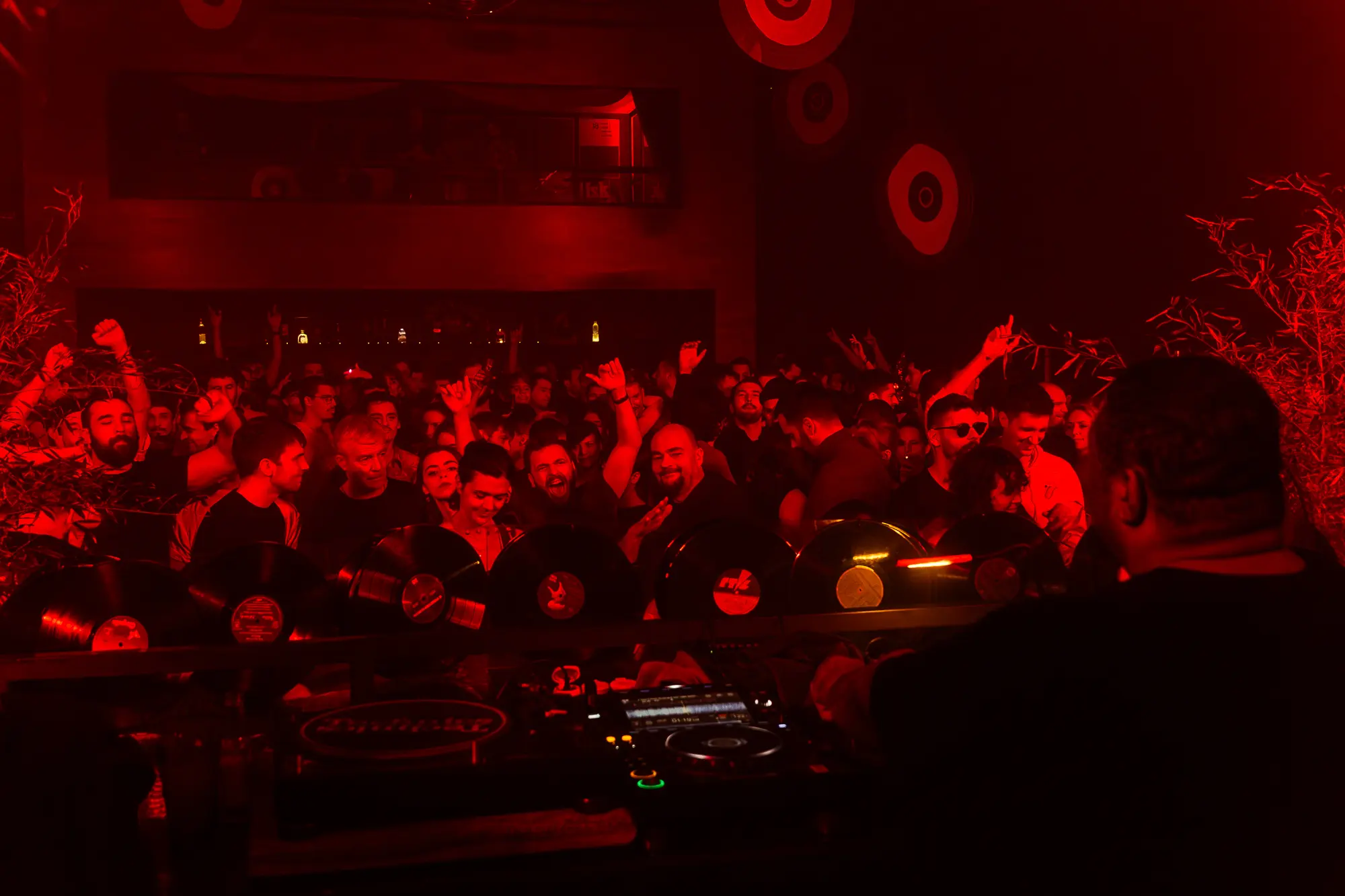 Crowd dancing in a red-lit nightclub with a DJ booth and vinyl records in the foreground.