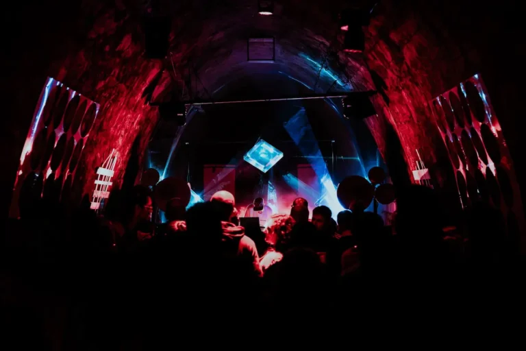 Crowd of people in a dark, cave-like club with red and blue lighting and a DJ booth on stage.