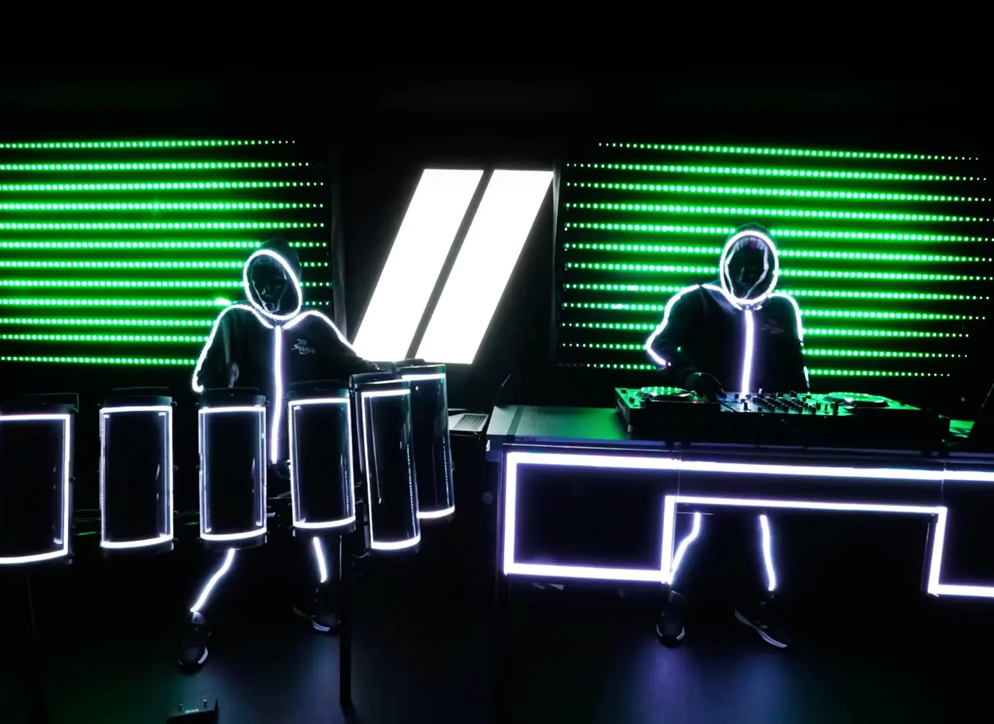 Two performers in glowing, neon-outlined suits perform at a dark stage, one at a DJ mixer and the other with lighted tubes.