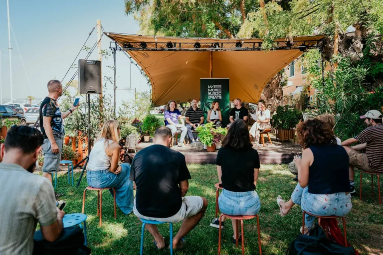 Outdoor panel discussion on a small stage with a tan canopy and a We Move Music Croatia banner, surrounded by a seated audience on colorful stools.