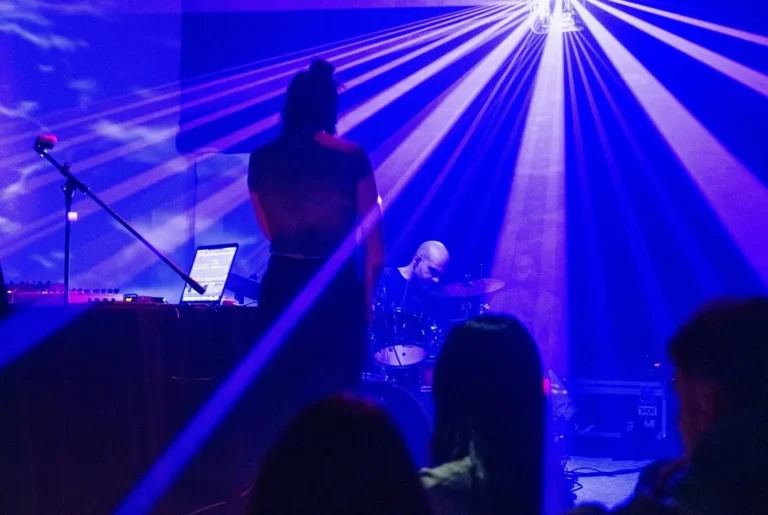 Musician performing on a stage under blue laser lights with a drummer in the background and a laptop on a stand to the left.