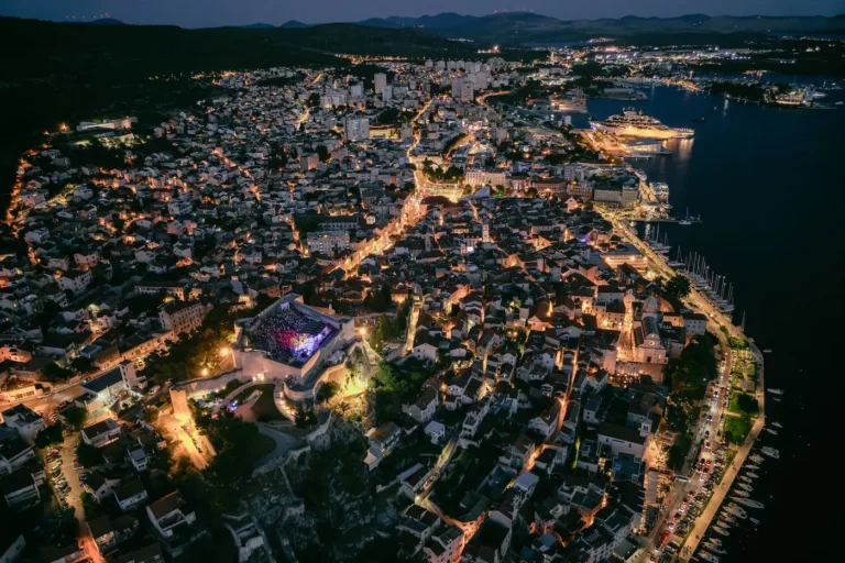 Aerial night view of a coastal town lit by streetlights, with a harbor and cruise ships on the right