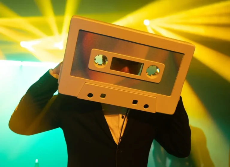 Person in a suit wears a oversized cassette tape as a headpiece on a neon-lit stage.