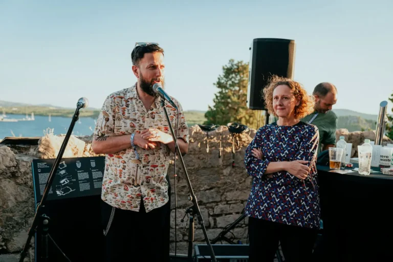 Two speakers on an outdoor stage by a stone wall, microphone in use, with a scenic sea backdrop.