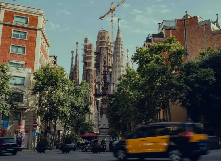 Street view in Barcelona with the Sagrada Família towers rising behind trees and a construction crane in the background, cars and taxis passing by.