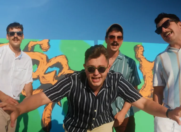 Four men posing in front of a bright mural, two wearing sunglasses, outdoors on a sunny day.