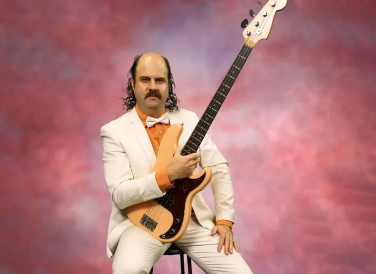 Man in a white suit with an orange shirt and bow tie holding a bass guitar against a pink backdrop.