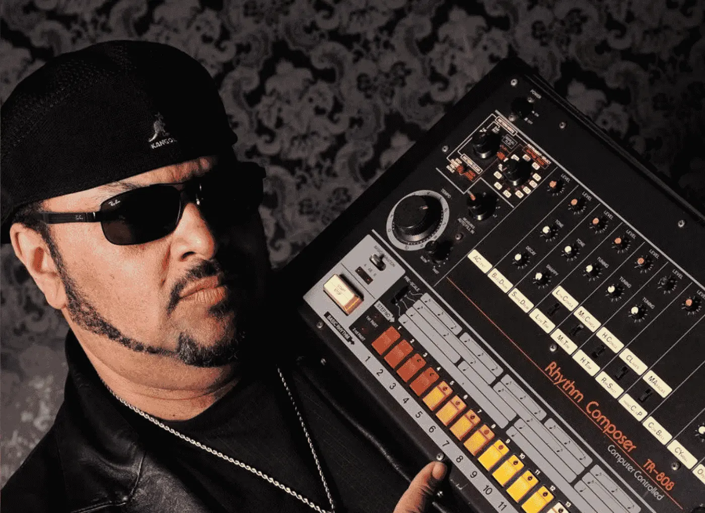 Musician in a black cap and sunglasses posing with a Roland TR-808 drum machine in a dim scene.