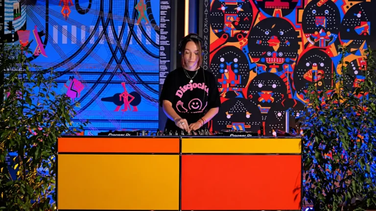 DJ wearing a black graphic tee mixes music at a bright orange-yellow-red console, with a vivid mural and plants framing the scene.