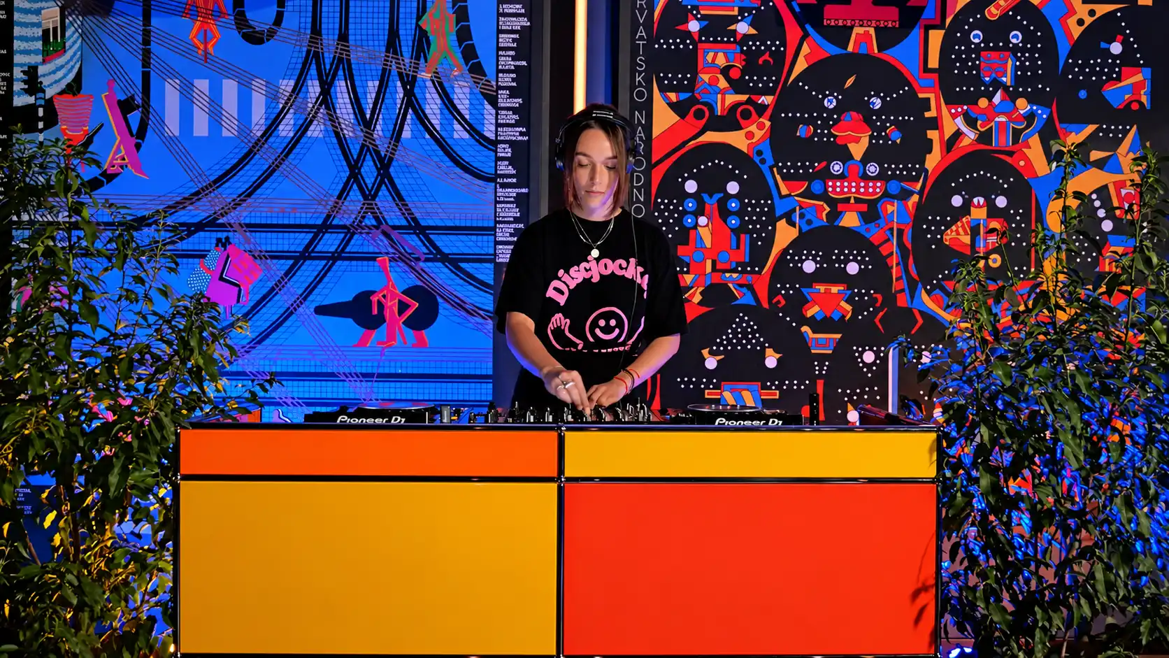 DJ wearing a black graphic tee mixes music at a bright orange-yellow-red console, with a vivid mural and plants framing the scene.