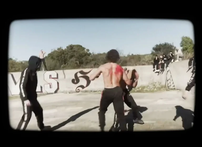 Shirtless man with red paint on his back fights another man in an outdoor concrete area; onlookers sit on a graffiti-covered wall.