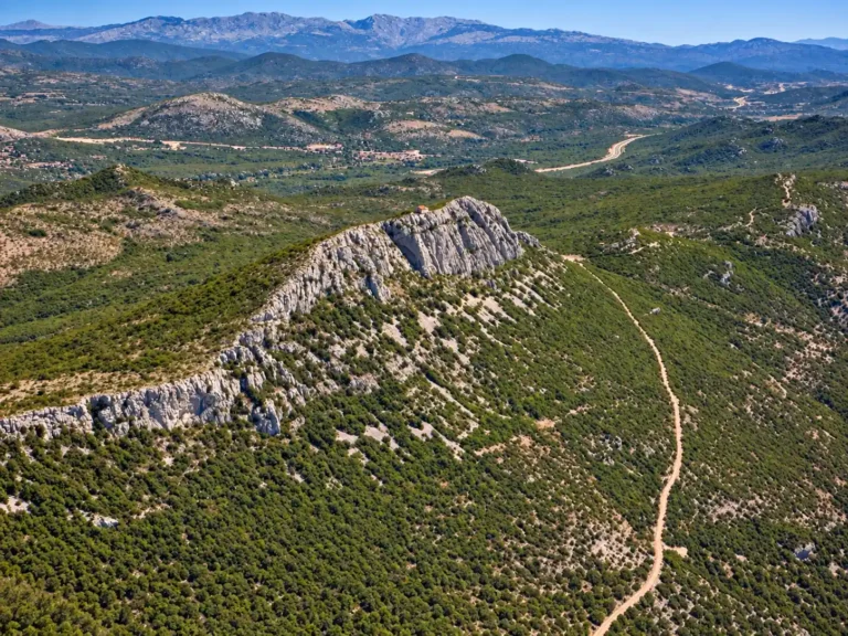 Aerial view of a white limestone ridge with green scrub and a winding dirt road through rolling hills toward distant mountains.