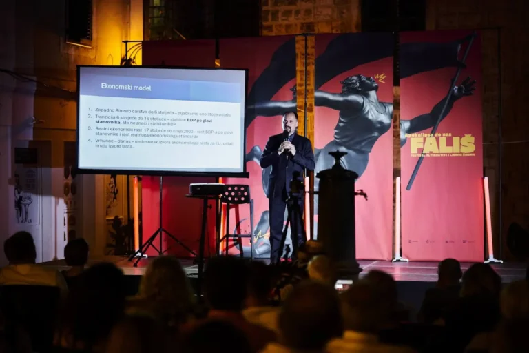 Male presenter on stage delivering a talk beside a projection screen showing a slide titled 'Ekonommski model' with an audience in the foreground.