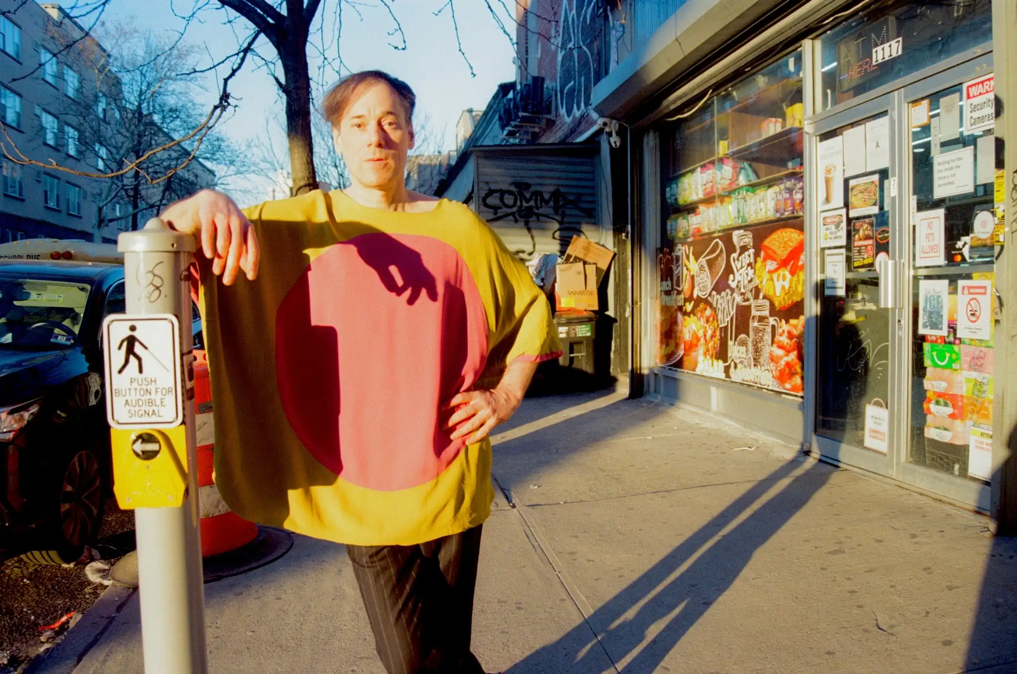 Man in a bright yellow shirt with a pink circular design leans on a pole on a city sidewalk beside a storefront covered in posters and graffiti on a sunny day.