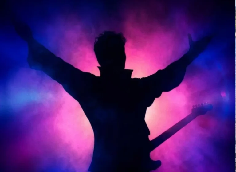 Silhouette of a guitarist with arms raised on a colorful purple-pink-blue stage backdrop.