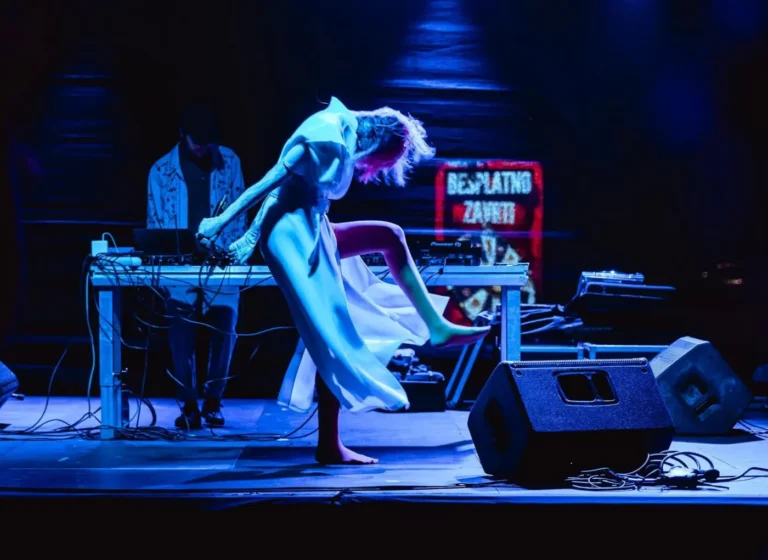 Dancer in a flowing white dress barefoot on stage, mid‑kick under blue lights with a DJ at a mixing desk behind.