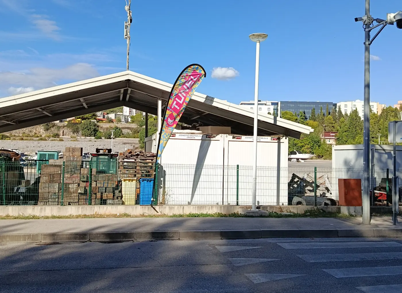 Outdoor industrial yard with pallets, containers, and a colorful curved banner near a metal shelter behind a fence.