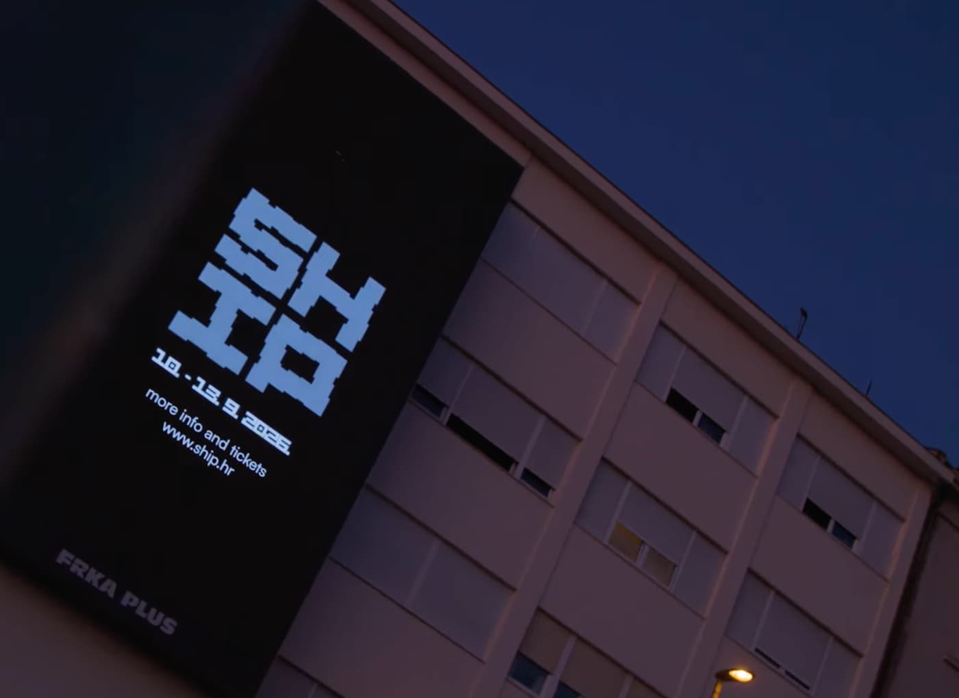 Night exterior of a building with a tall black banner displaying a bold white blocky logo and event text (dates and a site URL visible: www.ship.hr); the beige facade and windows of the building are dimly lit at dusk/night.