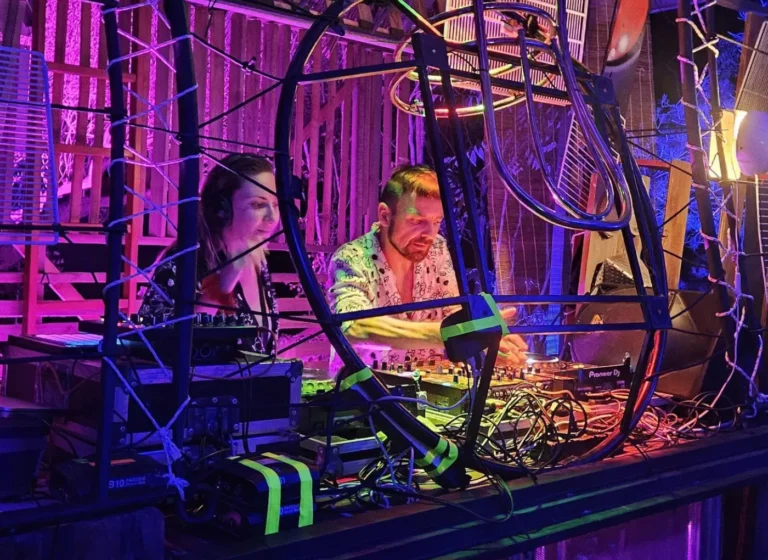 Two DJs in a neon-lit booth, surrounded by tangled cables and a metal frame, one man adjusting a Pioneer DJ mixer while a woman watches/assists nearby.