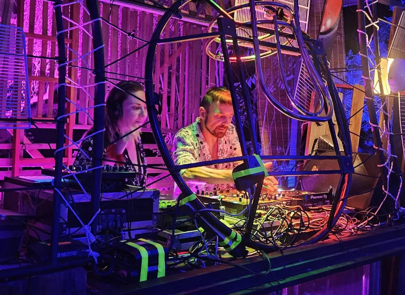 Two DJs in a neon-lit booth, surrounded by tangled cables and a metal frame, one man adjusting a Pioneer DJ mixer while a woman watches/assists nearby.