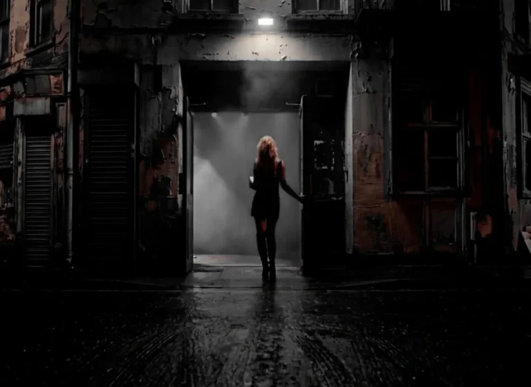 A woman in a dark coat stands in a dim doorway of a weathered building on a rainy street at night, with fog behind her.
