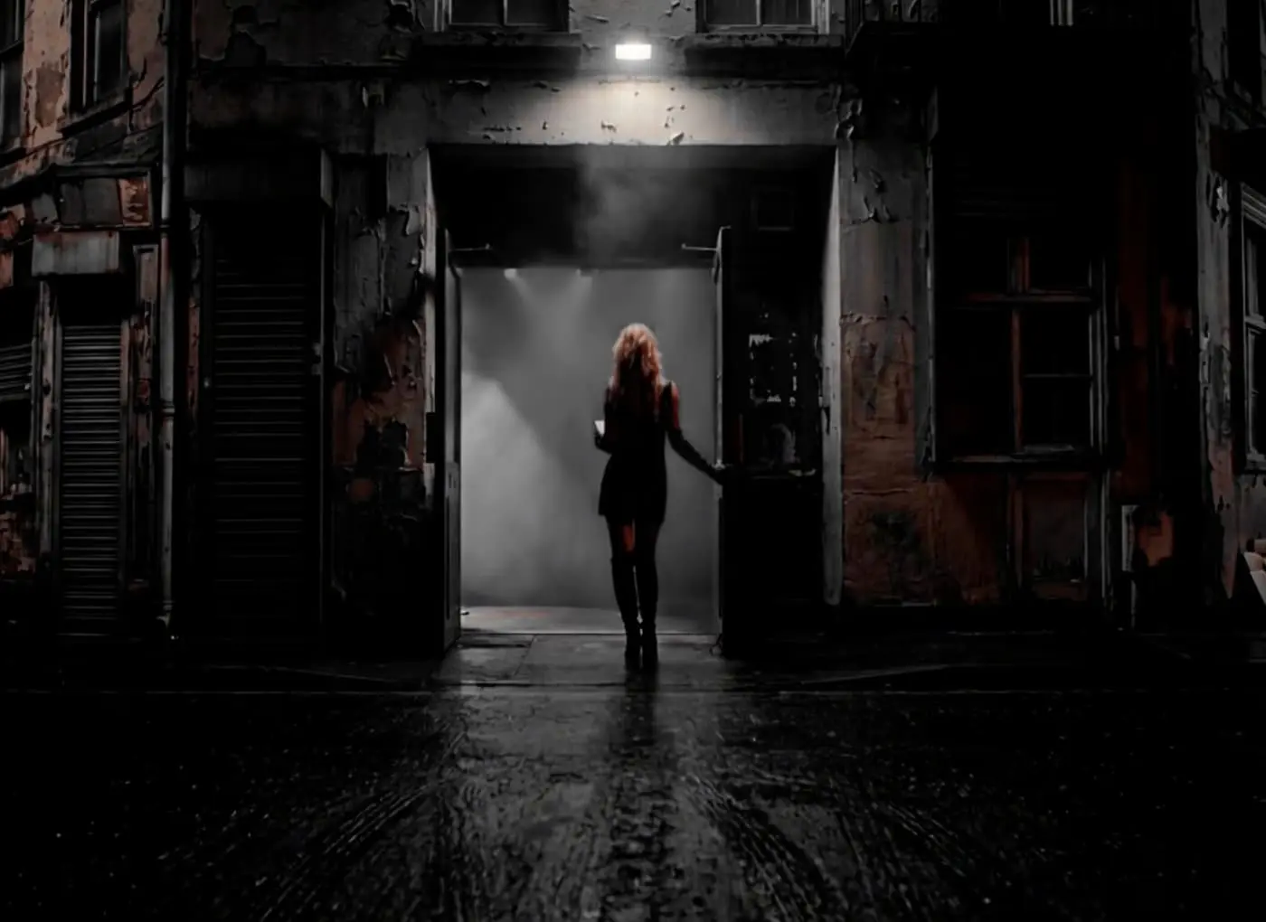 A woman in a dark coat stands in a dim doorway of a weathered building on a rainy street at night, with fog behind her.