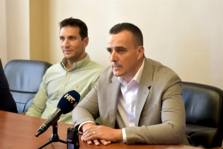 Two men sit at a conference table during a press briefing; the man on the right speaks into a microphone while the other listens.
