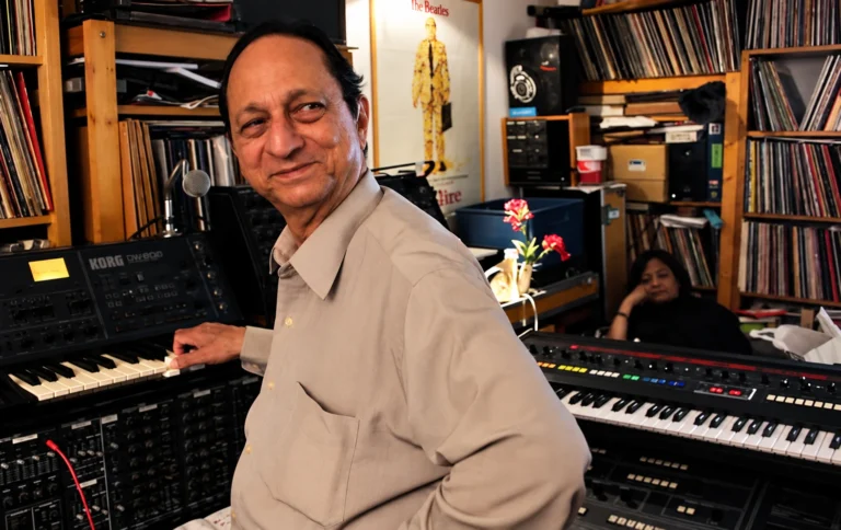 Man smiling while seated at a Korg synthesizer in a busy home studio with vinyl records on shelves behind him.
