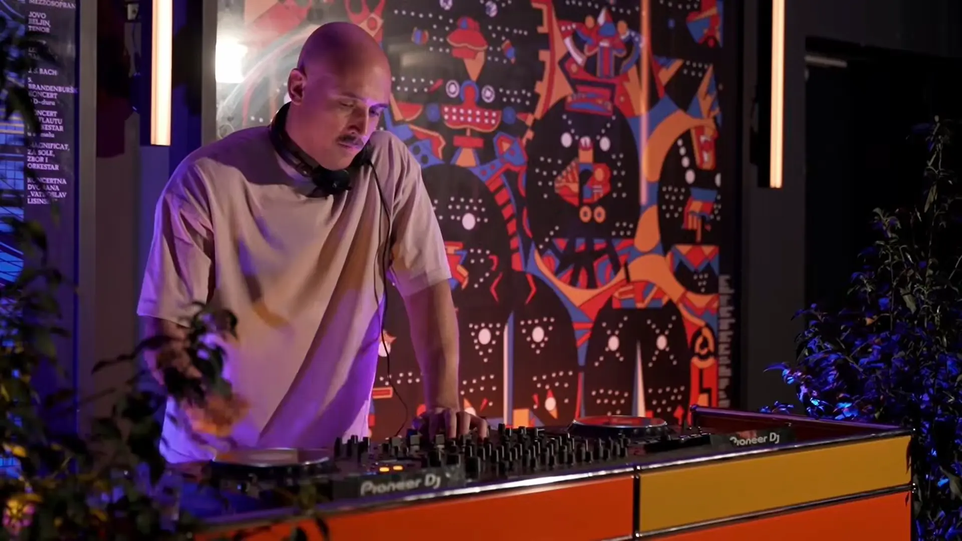 DJ in a beige shirt with headphones around his neck, mixing on a Pioneer DJ controller in a colorful mural-lit room behind him